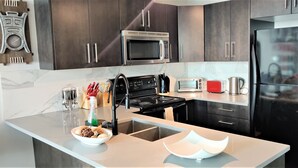 A modern kitchen with dark cabinets, stainless steel appliances, a marble countertop, and a central island with a sink. - Monki-Di Executive Suites - Alura (Calgary)