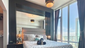 A modern bedroom with a large bed, a nightstand, a mirror, and a view of the city through large windows. - Monki-Di Executive Suites - Alura (Calgary)