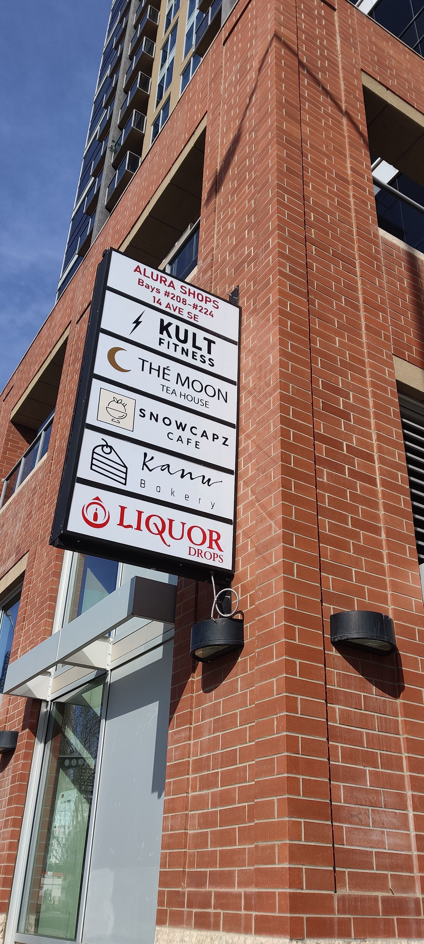 A building with a sign listing businesses including Alura Shops, Kult Fitness, The Moon Tea House, Snowca Cafe, Kanwa Bakery, and Liquor Drops.
