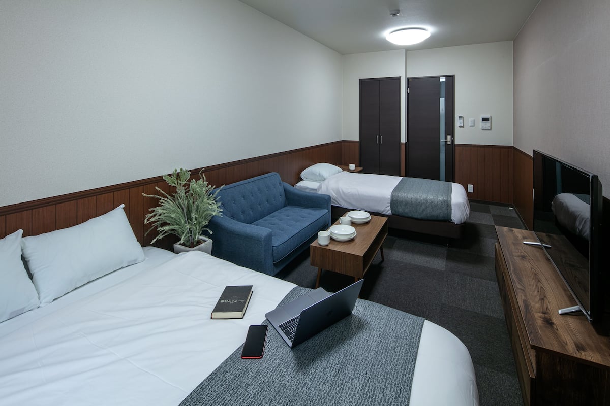 Une chambre d’hôtel avec un lit, un canapé, un bureau, un ordinateur portable, un téléphone portable et une plante en pot.