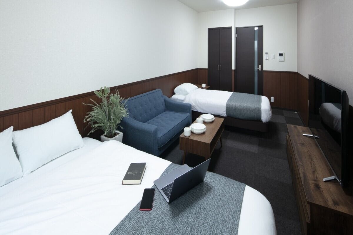 A hotel room with a bed, a desk with a laptop, a chair, a sofa, a TV, and a wardrobe.
