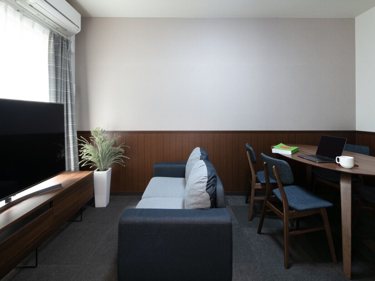 Moderni hotellihuone, jossa on taulutelevisio, sohva, ruokapöytä tuoleineen ja ruukkukasvi.