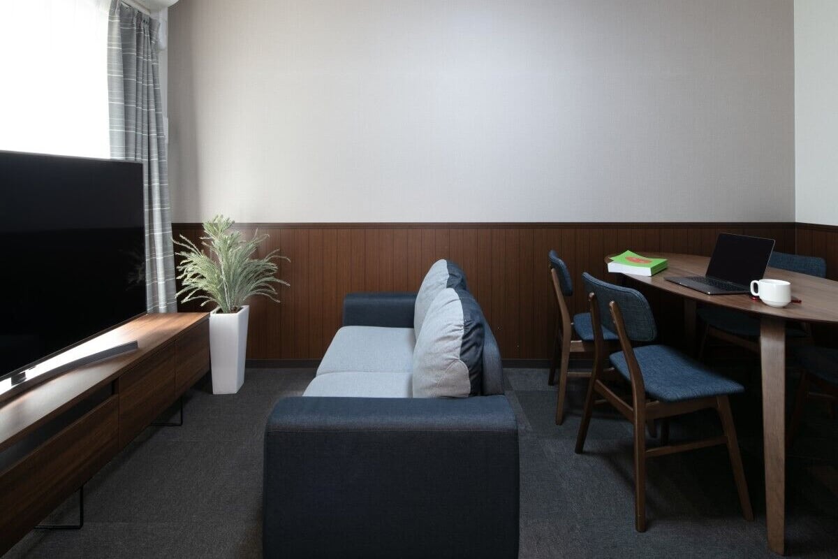 A modern hotel room with a flat-screen TV, a sofa, a dining table with chairs, and a potted plant.