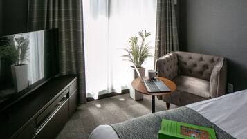 A hotel room with a bed, a desk with a laptop, a television, a potted plant, and a window with curtains.