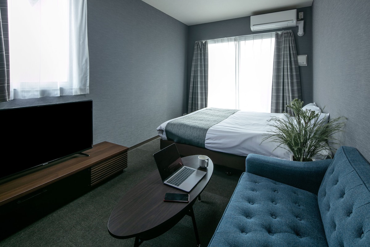 Une chambre d’hôtel avec un lit, une télévision, un ordinateur portable, une tasse à café, une plante et un canapé bleu.