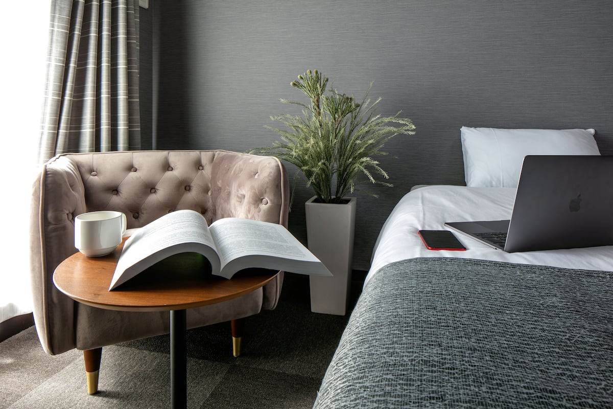 Une chambre d’hôtel avec un fauteuil à capitons, une petite table ronde sur laquelle se trouvent un livre et une tasse, une plante en pot, un lit avec un ordinateur portable et un téléphone intelligent posé sur le lit.