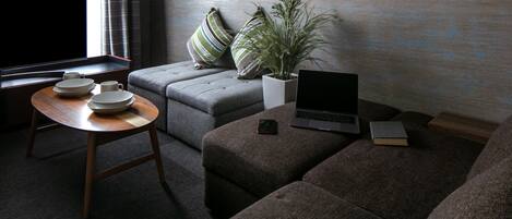 Una sala de estar moderna con un sofá gris, una mesita de madera con platos y una taza, una computadora portátil y una planta en maceta.