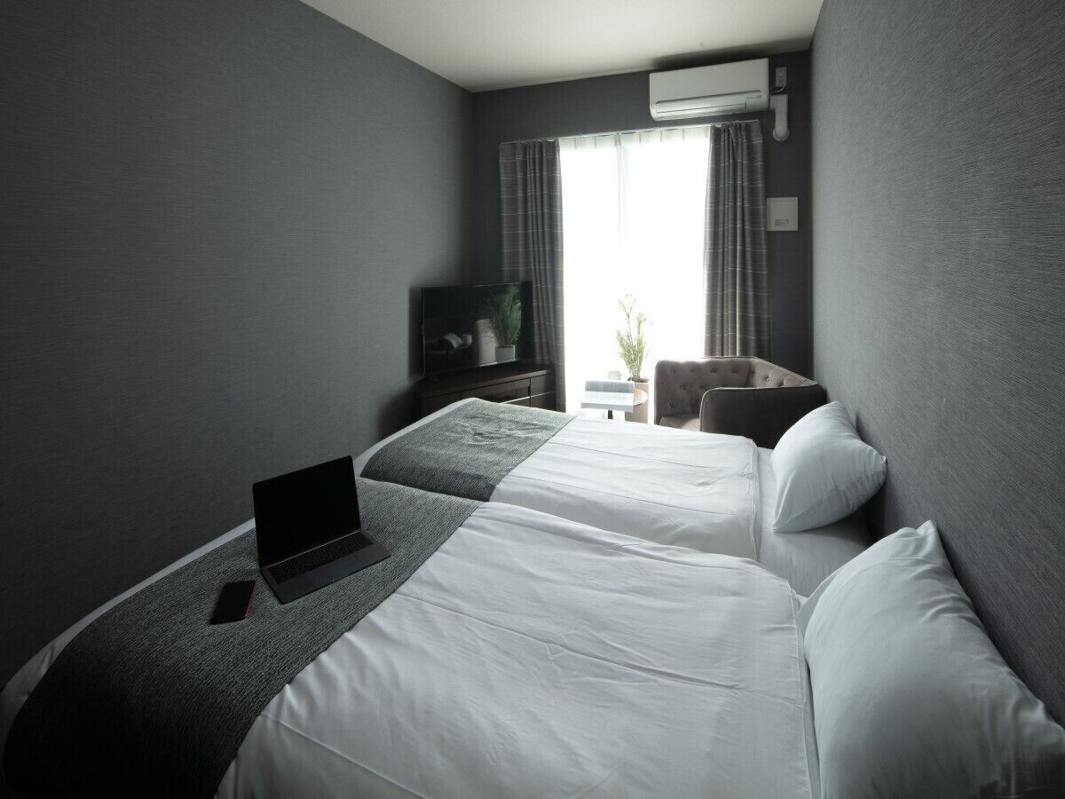 Een hotelkamer met twee bedden, een laptop op het linkerbed, een televisie aan de muur en een raam met gordijnen.