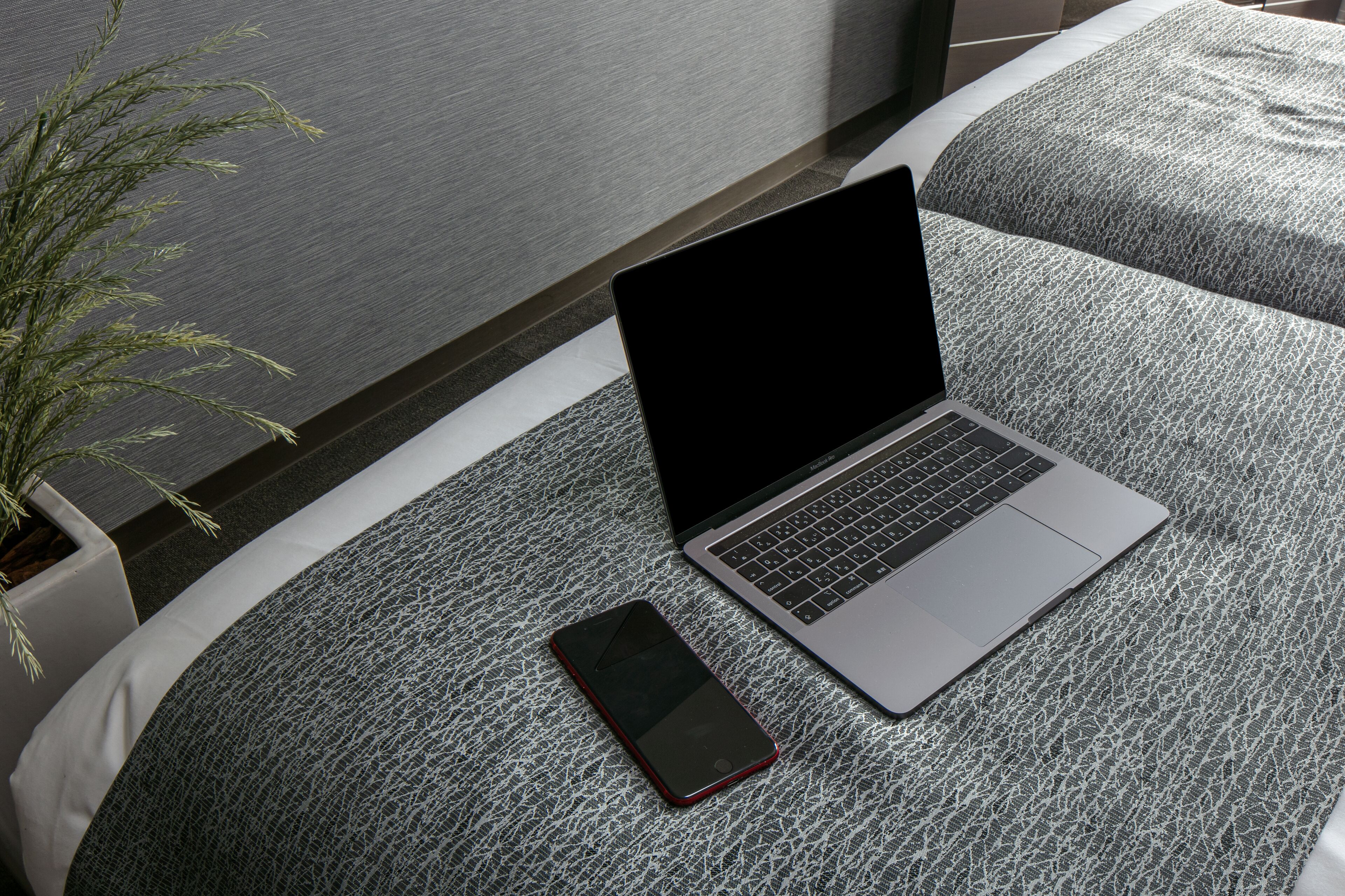 A laptop and a smartphone on a textured bed with a potted plant beside it.