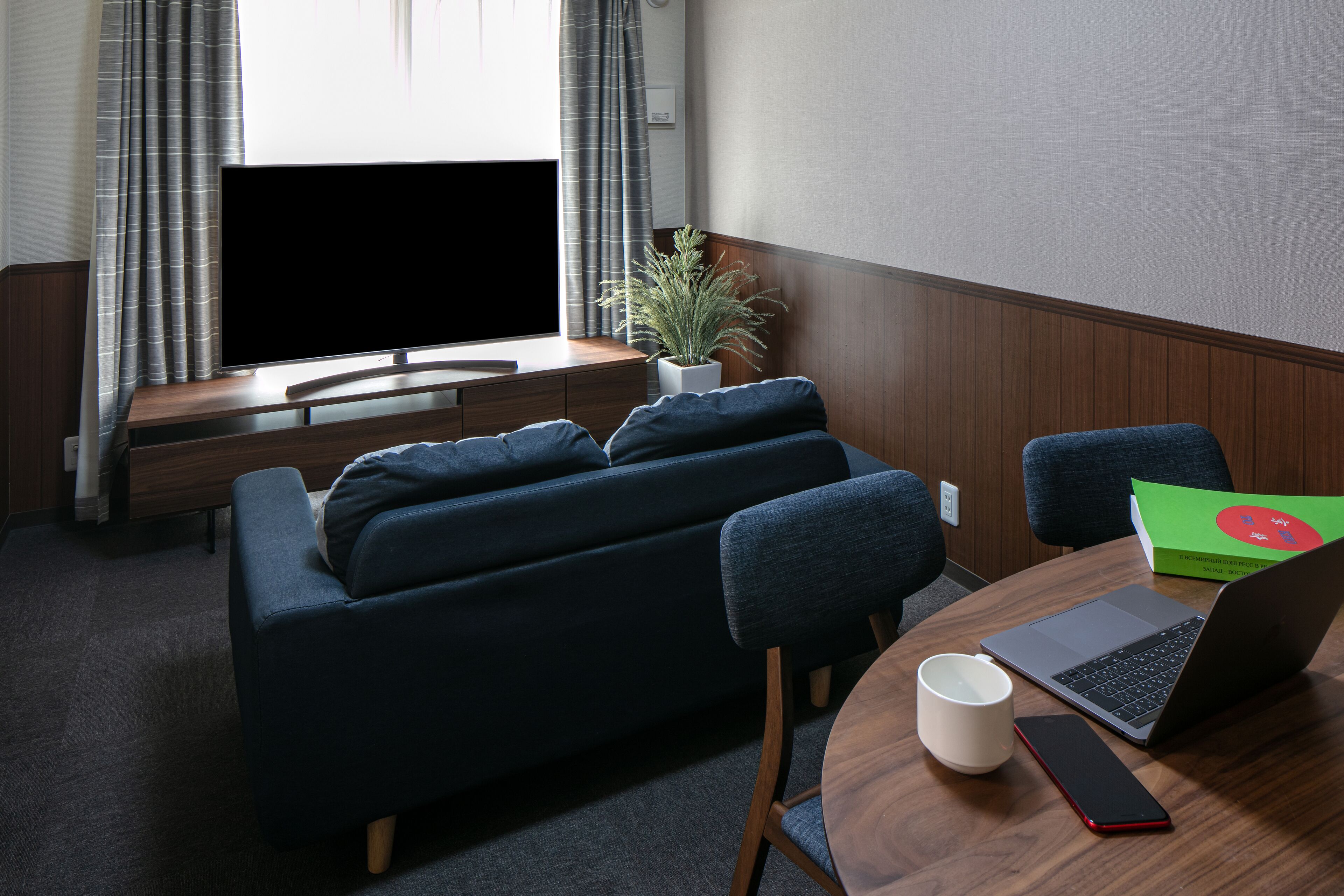 A modern living room with a dark blue sofa, a wooden coffee table, a laptop, a smartphone, and a potted plant.