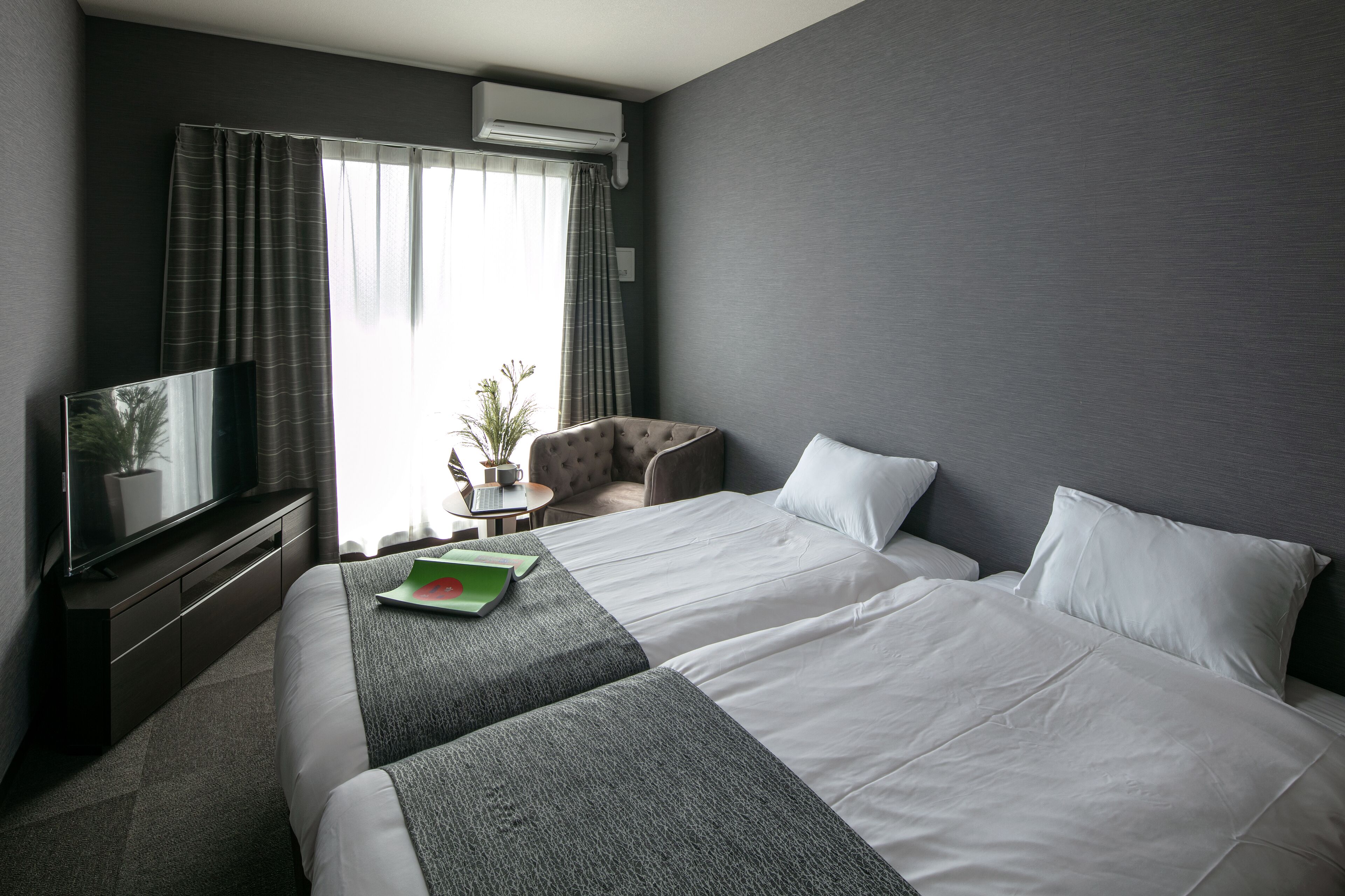 A hotel room with two beds, a flat-screen TV, a small table with a plant, and a window with curtains.