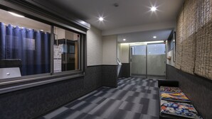 Un couloir d’hôtel avec un sol à carreaux, un comptoir de réception, un coin salon avec des coussins à motifs et une porte en verre donnant sur une chambre.