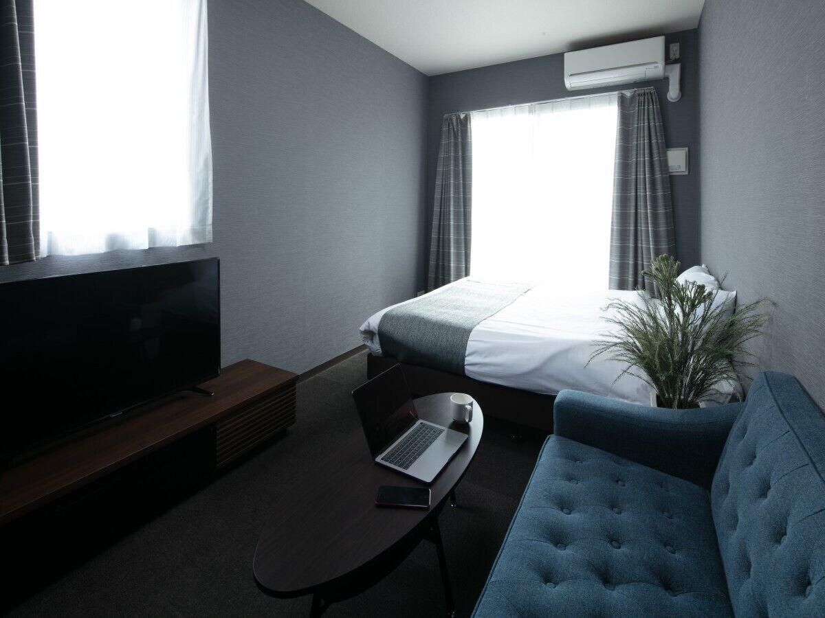 Hotelkamer met een bed, een televisie, een laptop, een koffiekop, een plant en een blauwe bank.