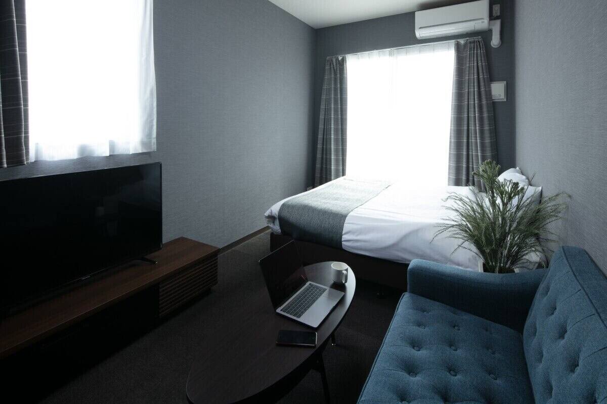 Une chambre d’hôtel avec un lit, une télévision, un ordinateur portable, une tasse à café, une plante et un canapé bleu.