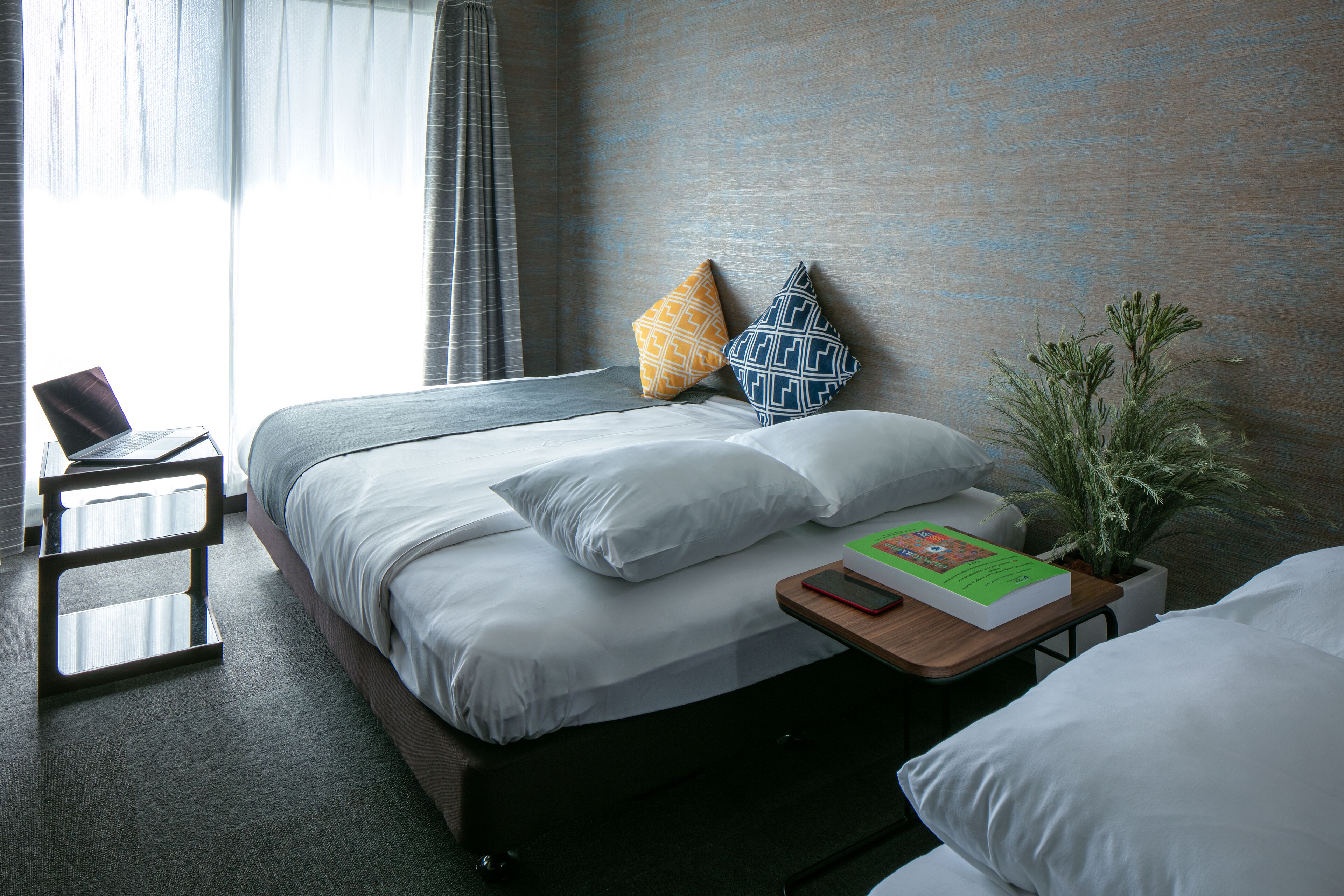 A neatly made bed with white linens, two patterned pillows, a bedside table with a book and a phone, and a potted plant on the floor.