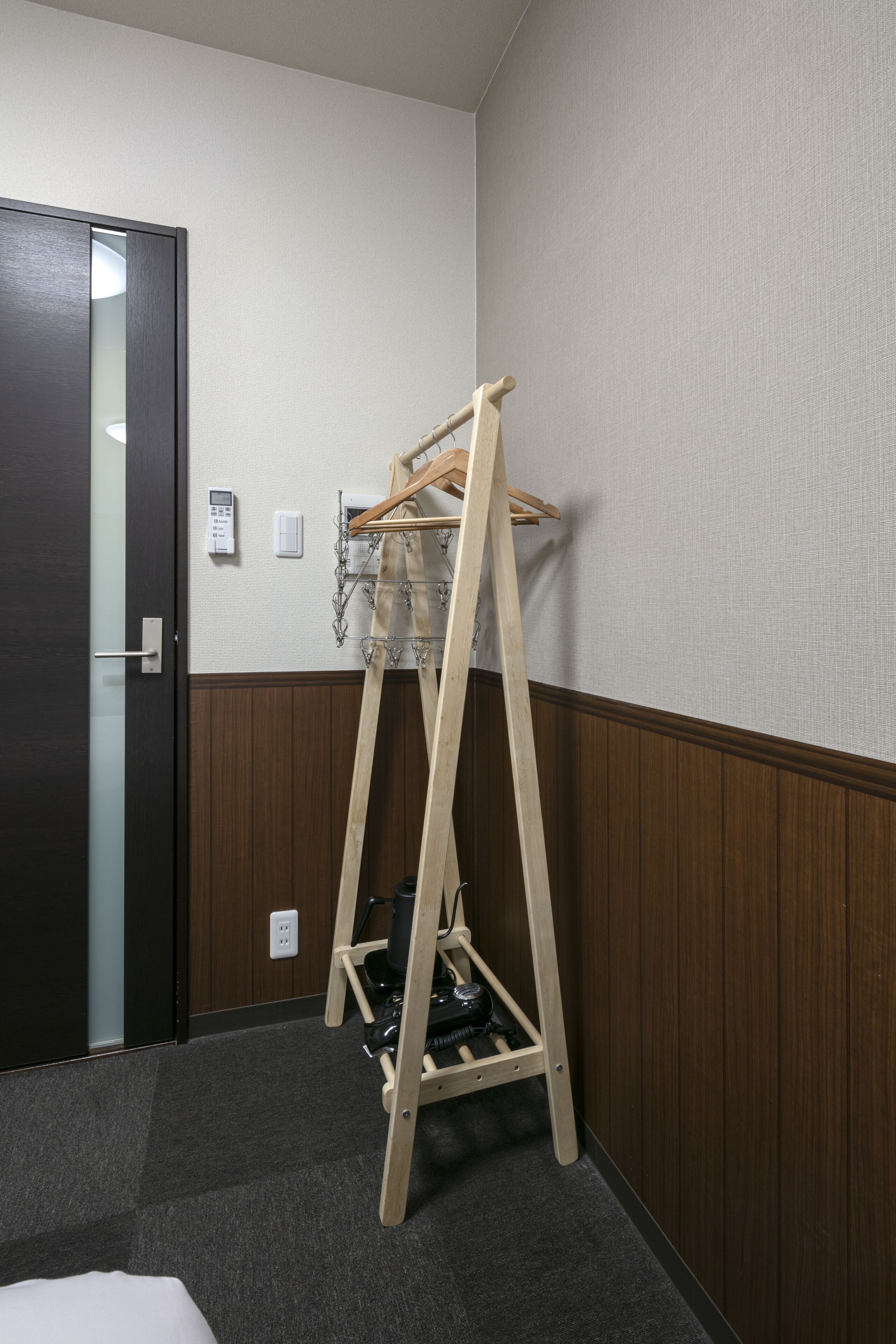 A corner of a hotel room with a wooden clothes rack, a door, and a wall-mounted thermostat.