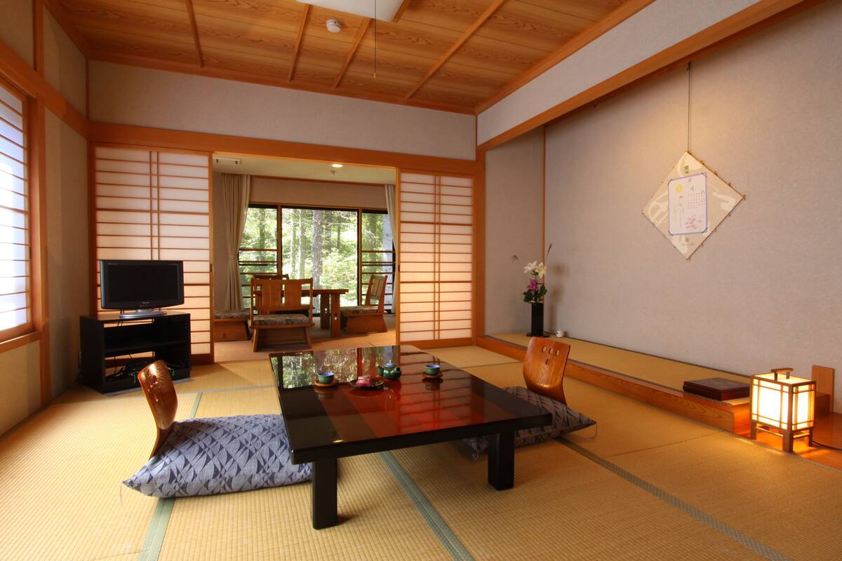 A traditional Japanese room with tatami flooring, a low table, cushions, a television, and a sliding door leading to a view of trees.