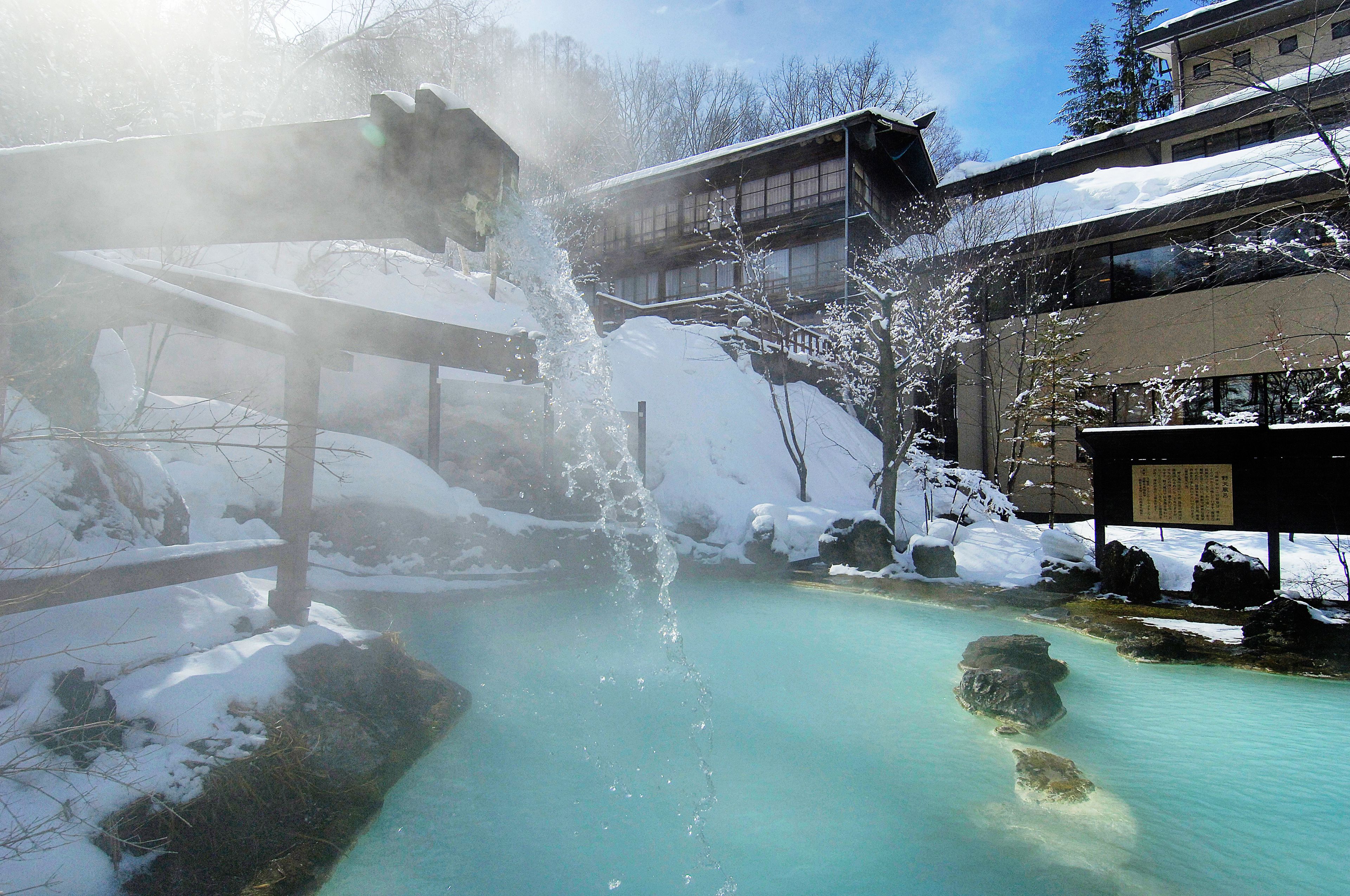 Een traditionele Japanse onsen met een houten looppad dat leidt naar een warmwaterbron, omgeven door met sneeuw bedekte rotsen en bomen.