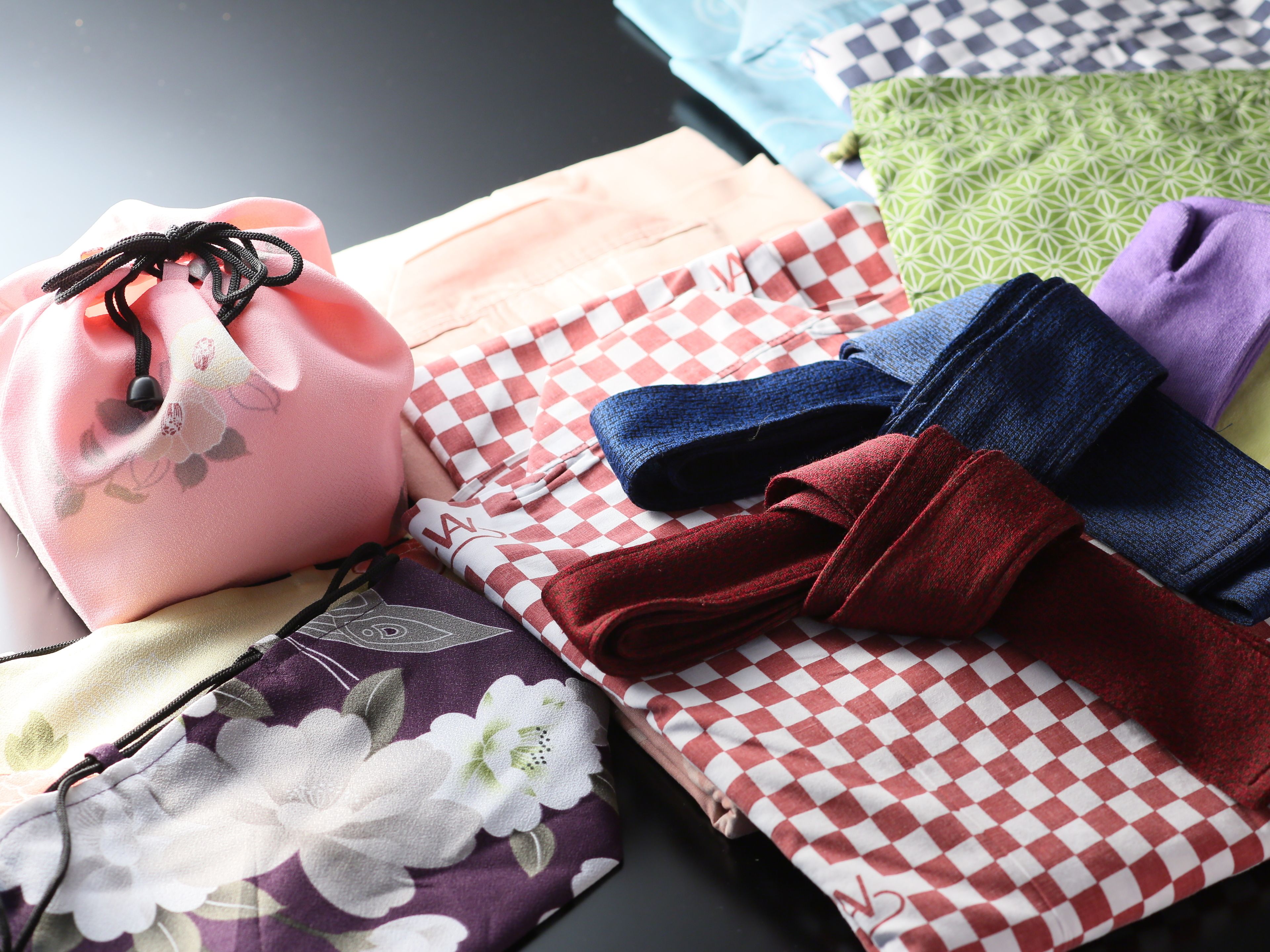 A variety of fabrics and textiles laid out on a table, including a pink fabric with a floral pattern, a red and white checkered fabric, and several other patterned fabrics in different colors.