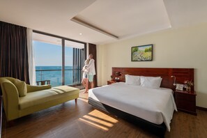 A hotel room with a large bed, a sofa, a nightstand with a lamp, a painting on the wall, and a view of the sea through a large window.