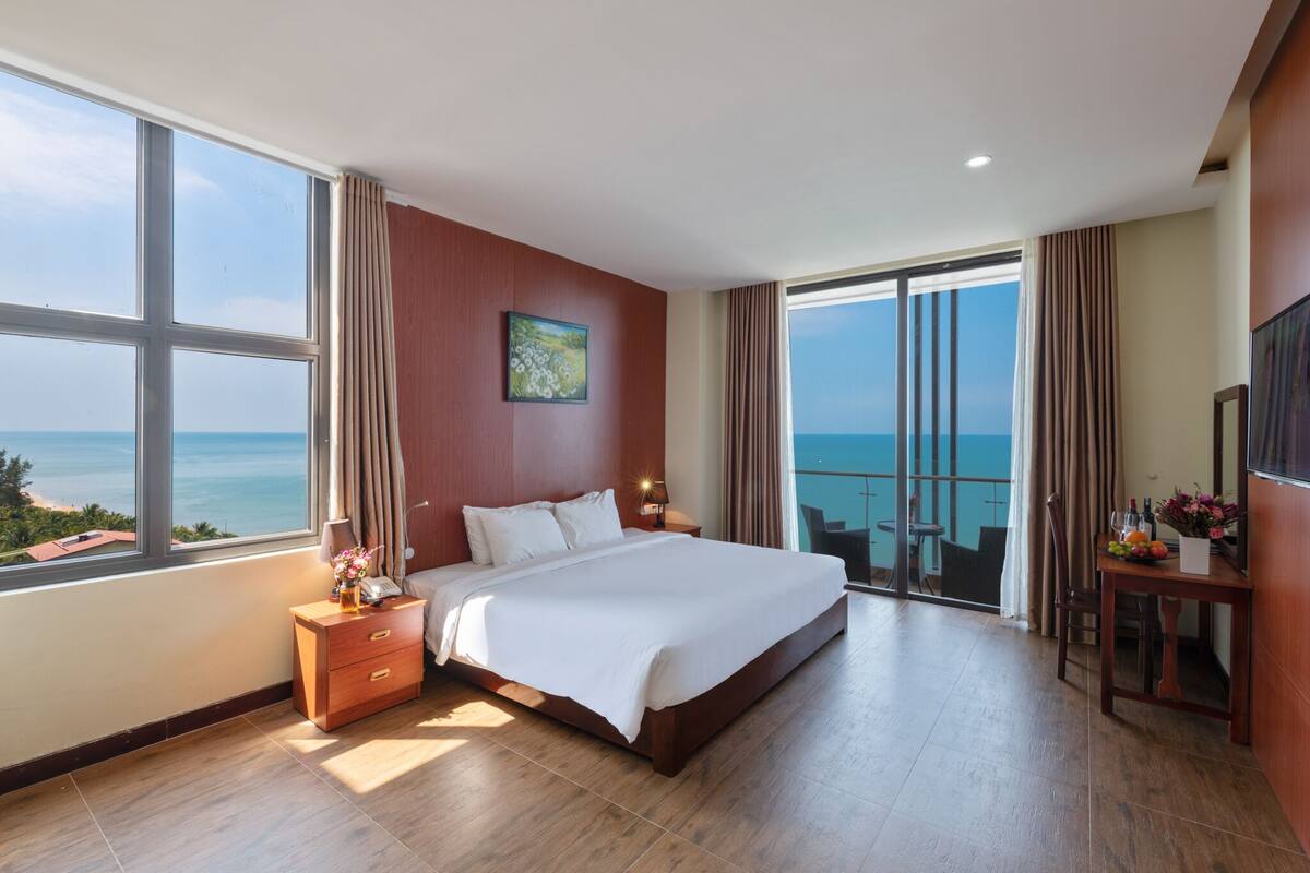 Une chambre spacieuse avec un grand lit, une table de chevet, un bureau avec une chaise et une vue sur l’océan grâce à de grandes fenêtres.
