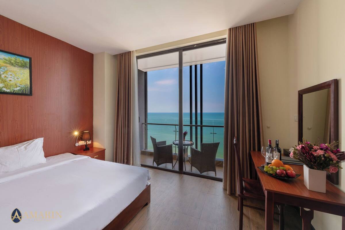 Une chambre d’hôtel avec un grand lit, un bureau agrémenté d’un vase de fleurs, une table avec des fruits et des bouteilles de vin, et un balcon offrant une vue sur la mer.
