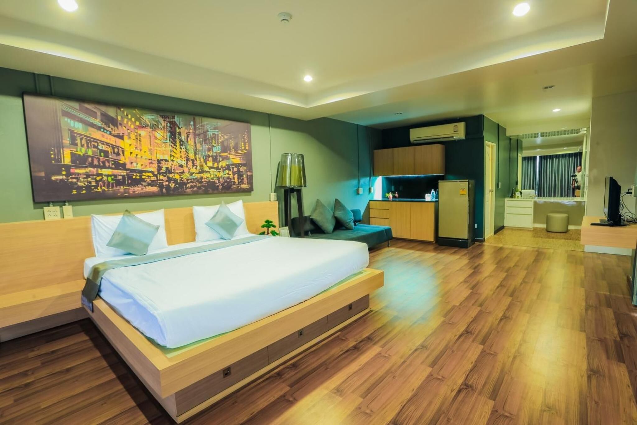 A modern hotel room with a large bed, wooden flooring, a kitchenette, and a cityscape painting on the wall.