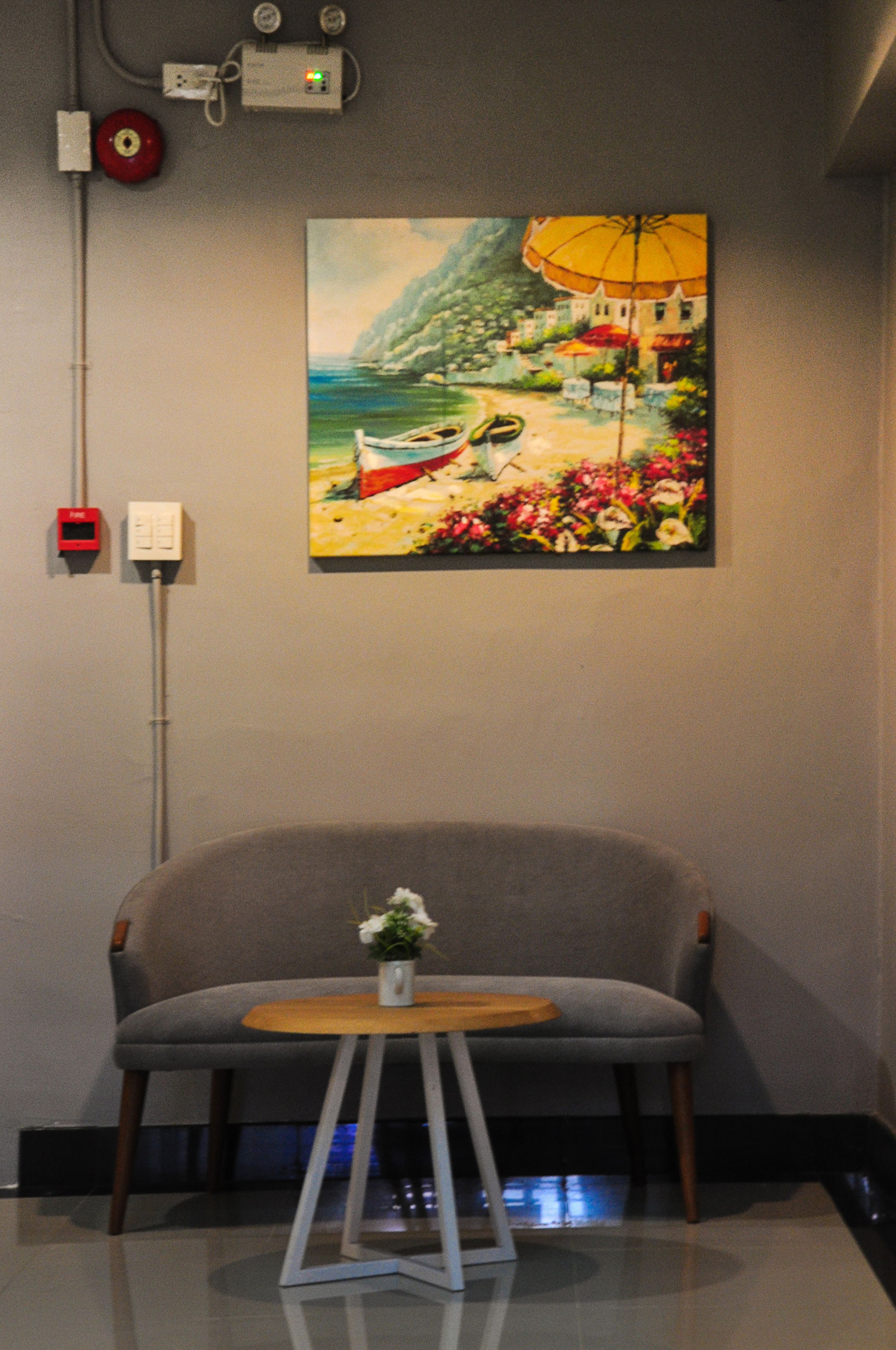 A room with a grey armchair, a small wooden table with a white vase, and a painting of a beach scene on the wall.