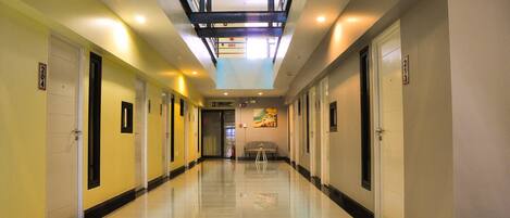 A long, well-lit hotel corridor with doors numbered 201 to 213, a central seating area, and a ceiling with a skylight.