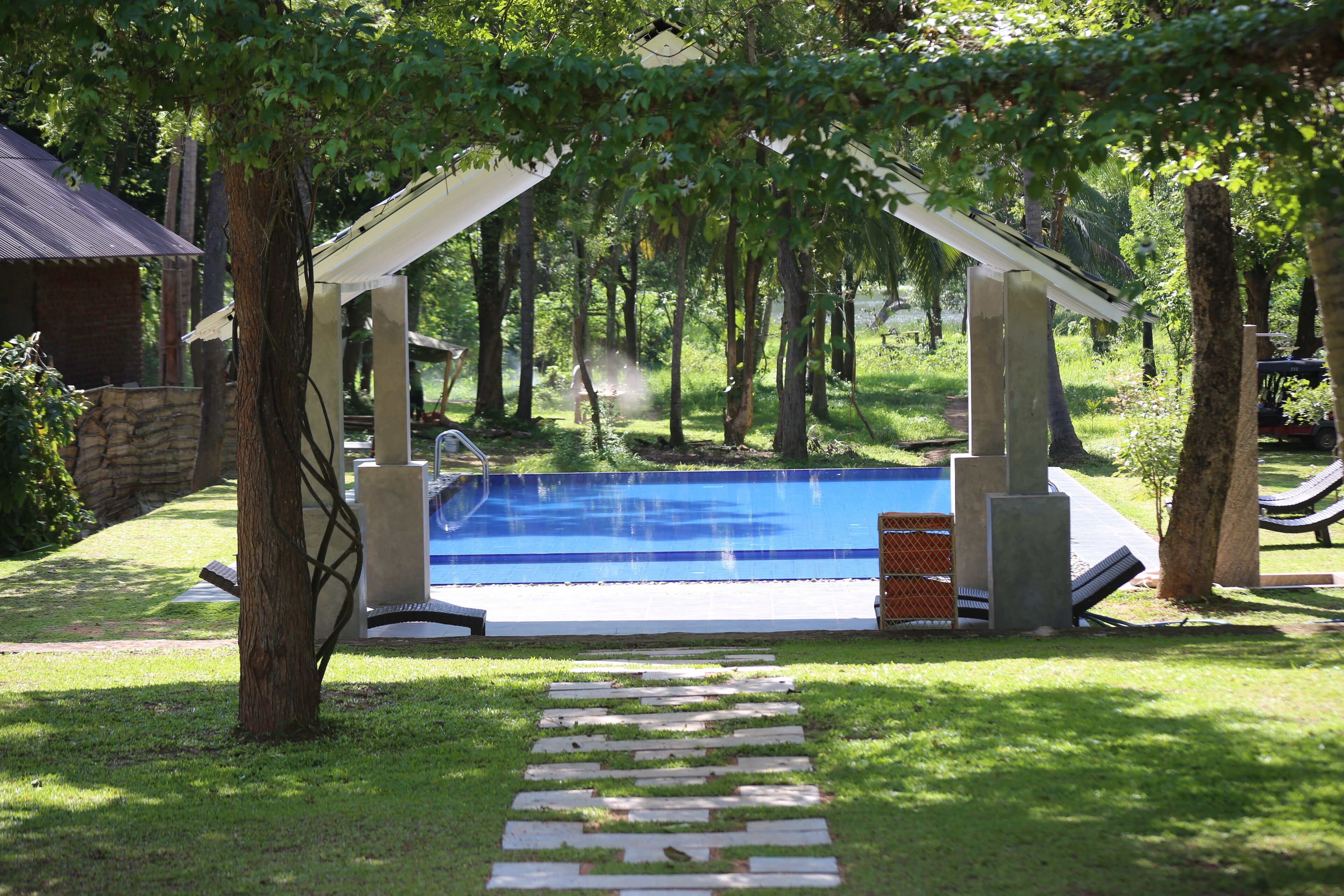 A swimming pool with a stone pathway leading to it, surrounded by trees and a covered area.