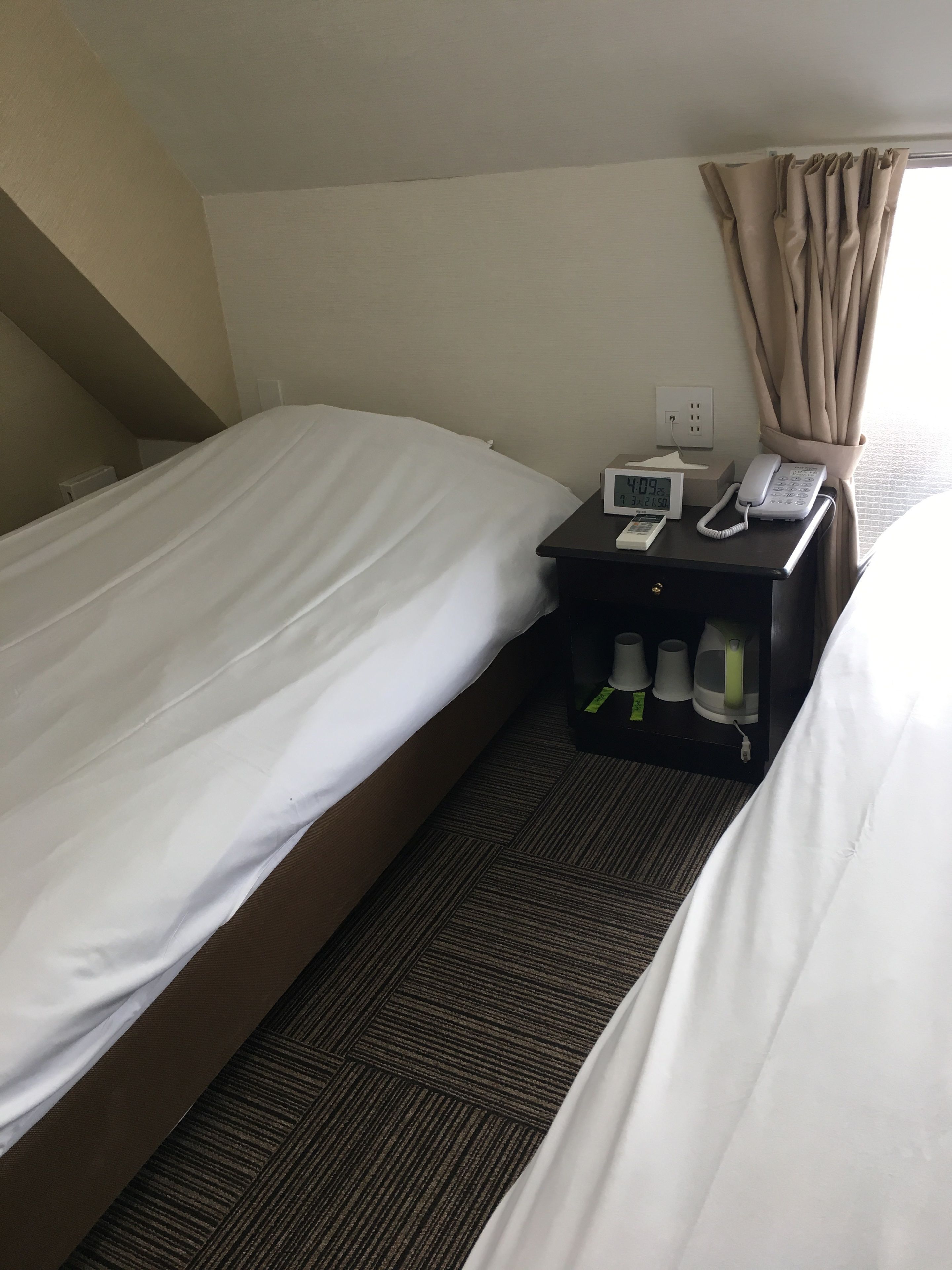 A hotel room with two beds, a bedside table with a phone and a clock, and a window with curtains.
