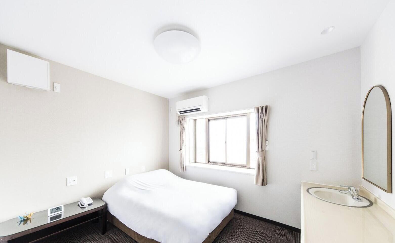 Une chambre d’hôtel comprenant un lit, une table de chevet avec un téléphone, une fenêtre avec des rideaux, un climatiseur fixé au mur et une salle de bain avec un lavabo et un miroir.