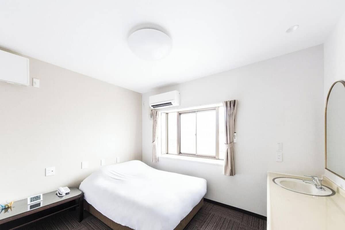 Une chambre d’hôtel comprenant un lit, une table de chevet avec un téléphone, une fenêtre avec des rideaux, un climatiseur fixé au mur et une salle de bain avec un lavabo et un miroir.