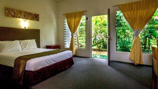 Habitación de hotel con cama, mesita de noche, lámpara y vista a zonas verdes a través de la puerta.