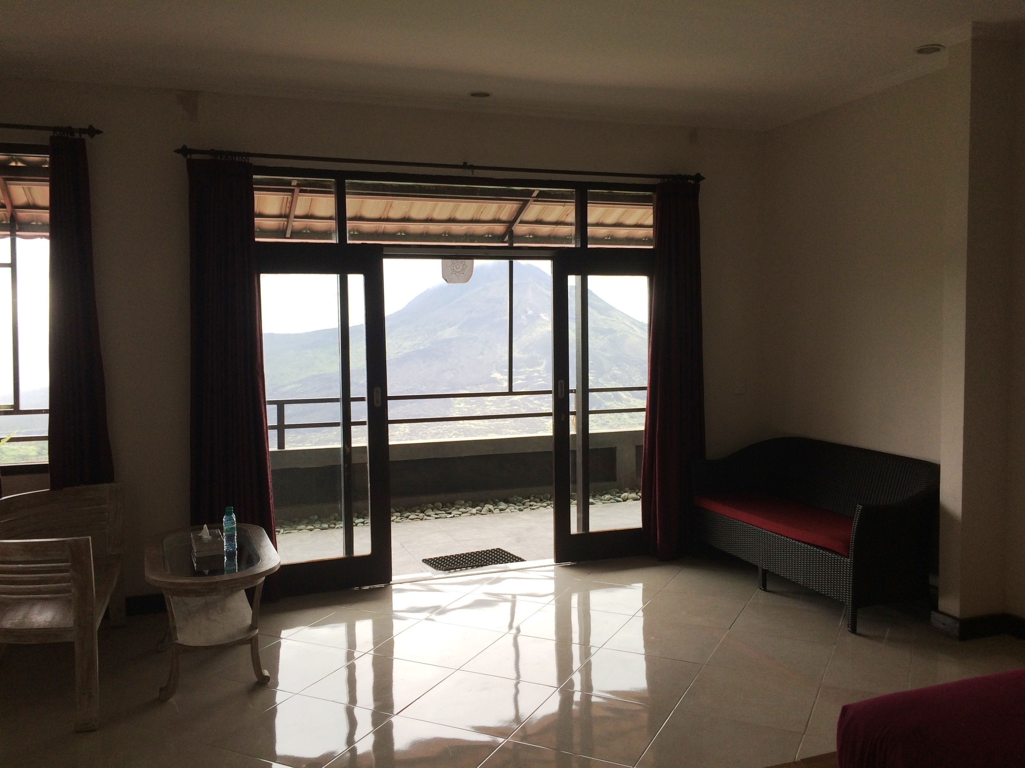 A room with a large sliding glass door leading to a balcony with a mountain view. There is a wooden chair, a small table with a bottle, and a red bench.
