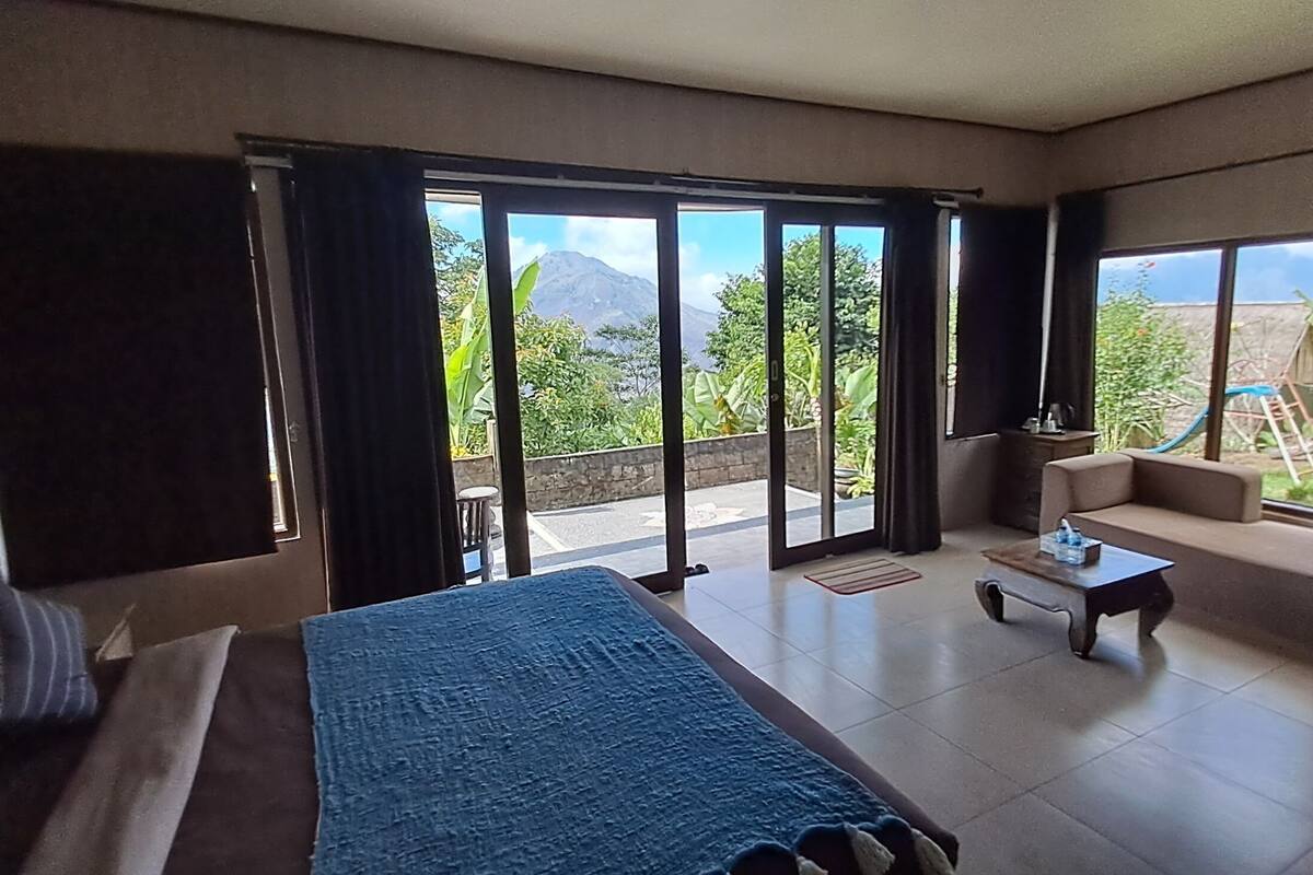 Quarto de hotel com cama, sofá, uma mesinha e vista para uma montanha através de grandes janelas.