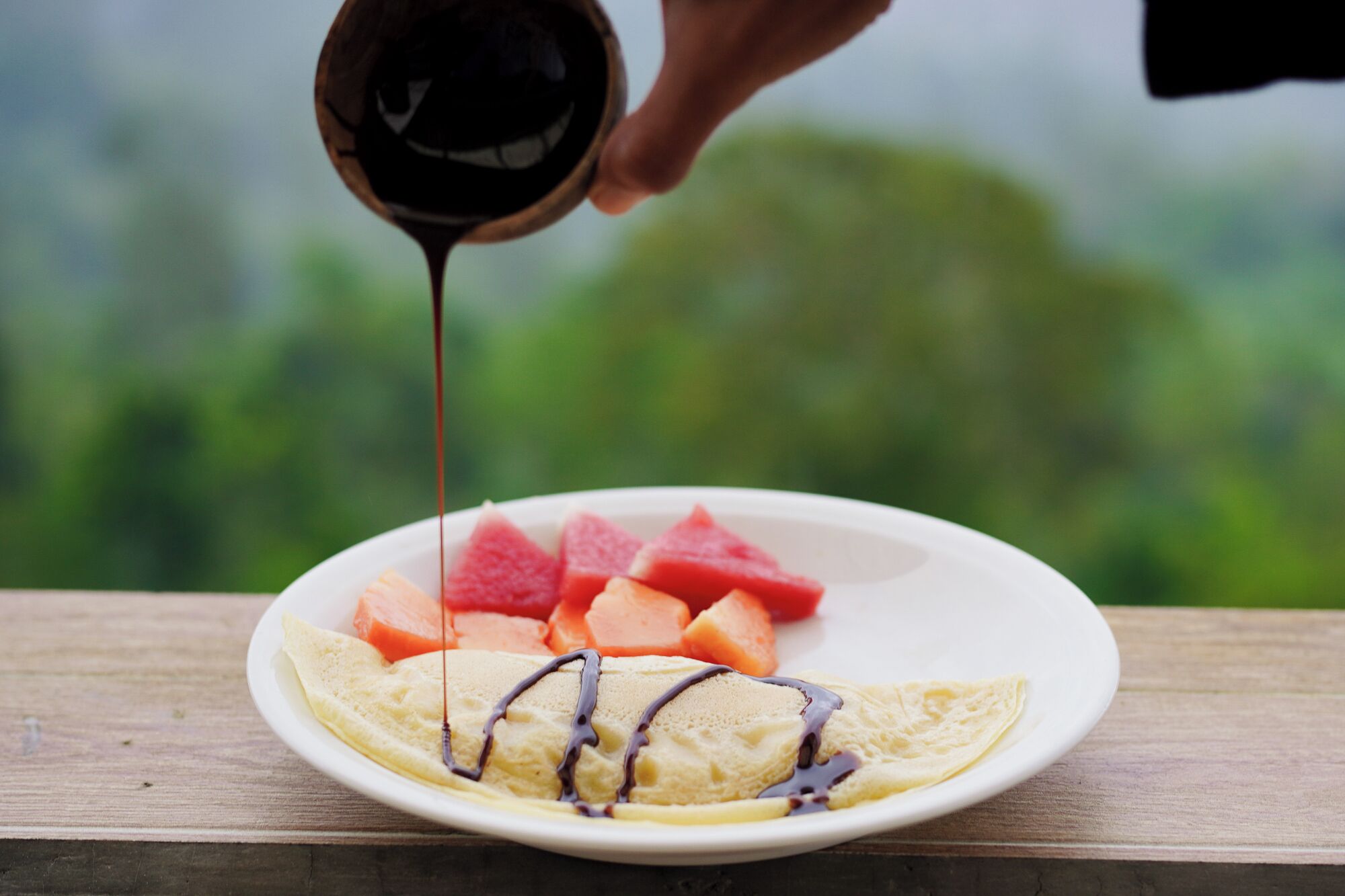 Ein Teller mit einer Crêpe, Wassermelone und Orangenscheiben, während Schokoladensauce darüber gegossen wird.