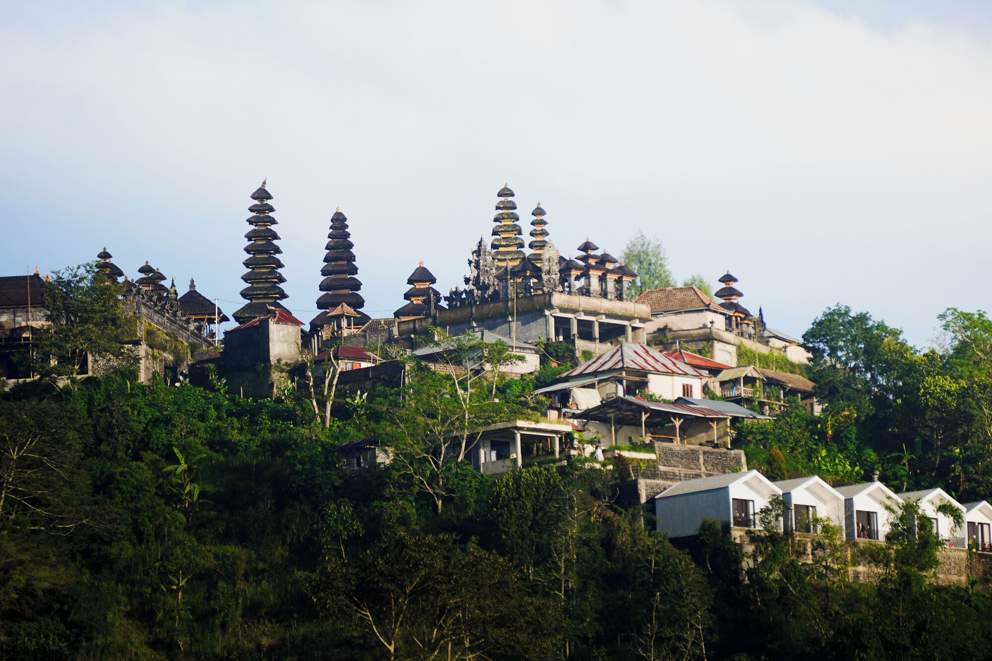 A traditional Balinese temple complex with tiered structures and ornate architecture, surrounded by lush greenery and residential buildings.
