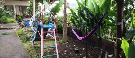 Un parque infantil con tobogán y estructura de escalada, rodeado de abundante vegetación y una hamaca.