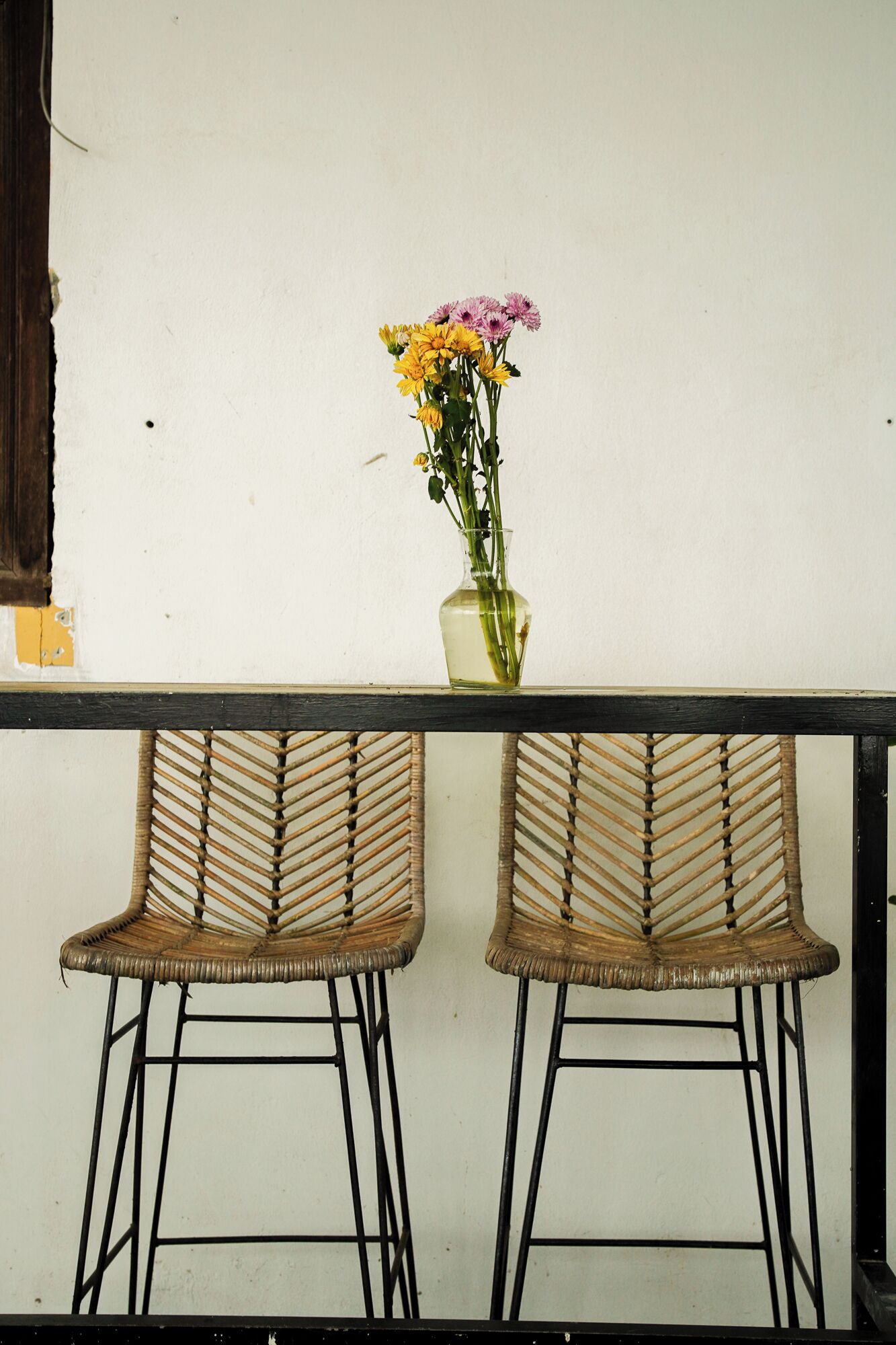 Two wicker chairs with metal frames, a glass vase with flowers, and a white wall.