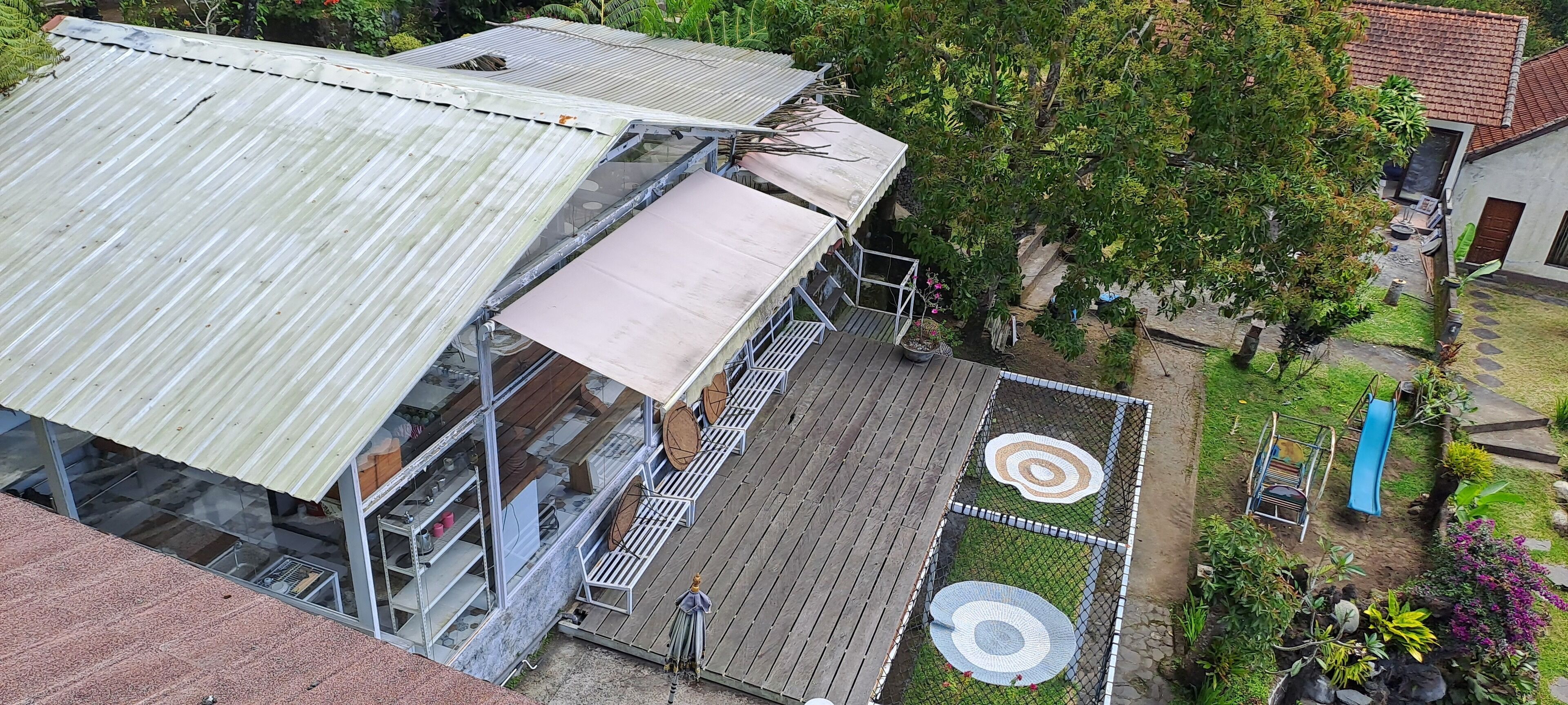 Een gebouw met een golfplaten dak, een houten terras met cirkelmotieven en een tuin met een glijbaan.