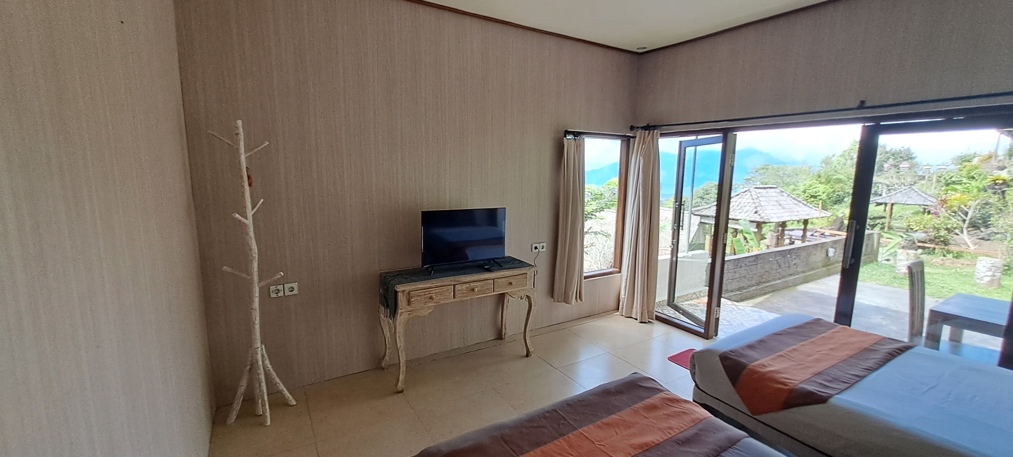 A bedroom with a bed, a desk with a television, and a sliding glass door leading to an outdoor area with a view of greenery and a thatched-roof structure.