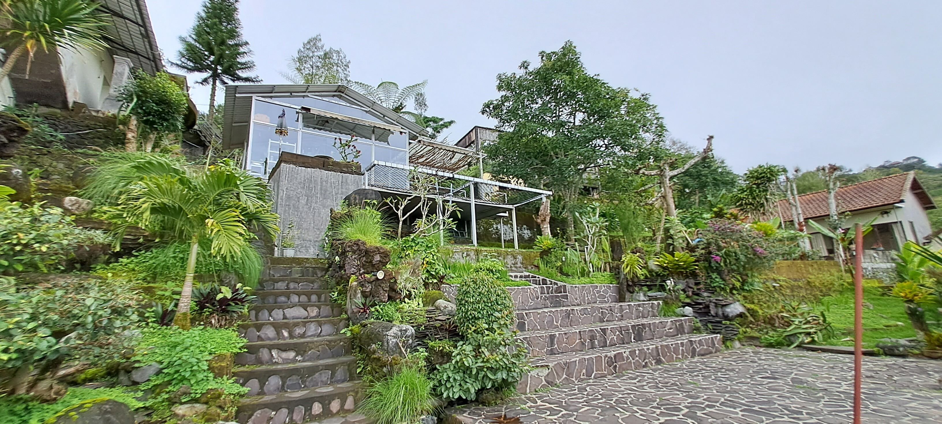 A stone staircase leading to a house with a glass balcony, surrounded by lush greenery and a hilly landscape.