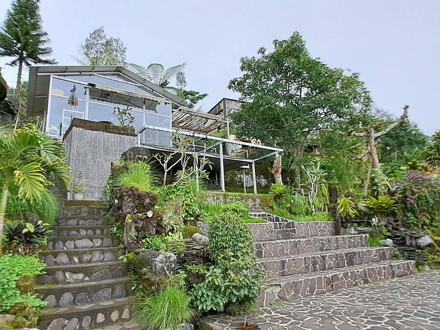 A stone staircase leading to a house with a glass balcony, surrounded by lush greenery and a hilly landscape.