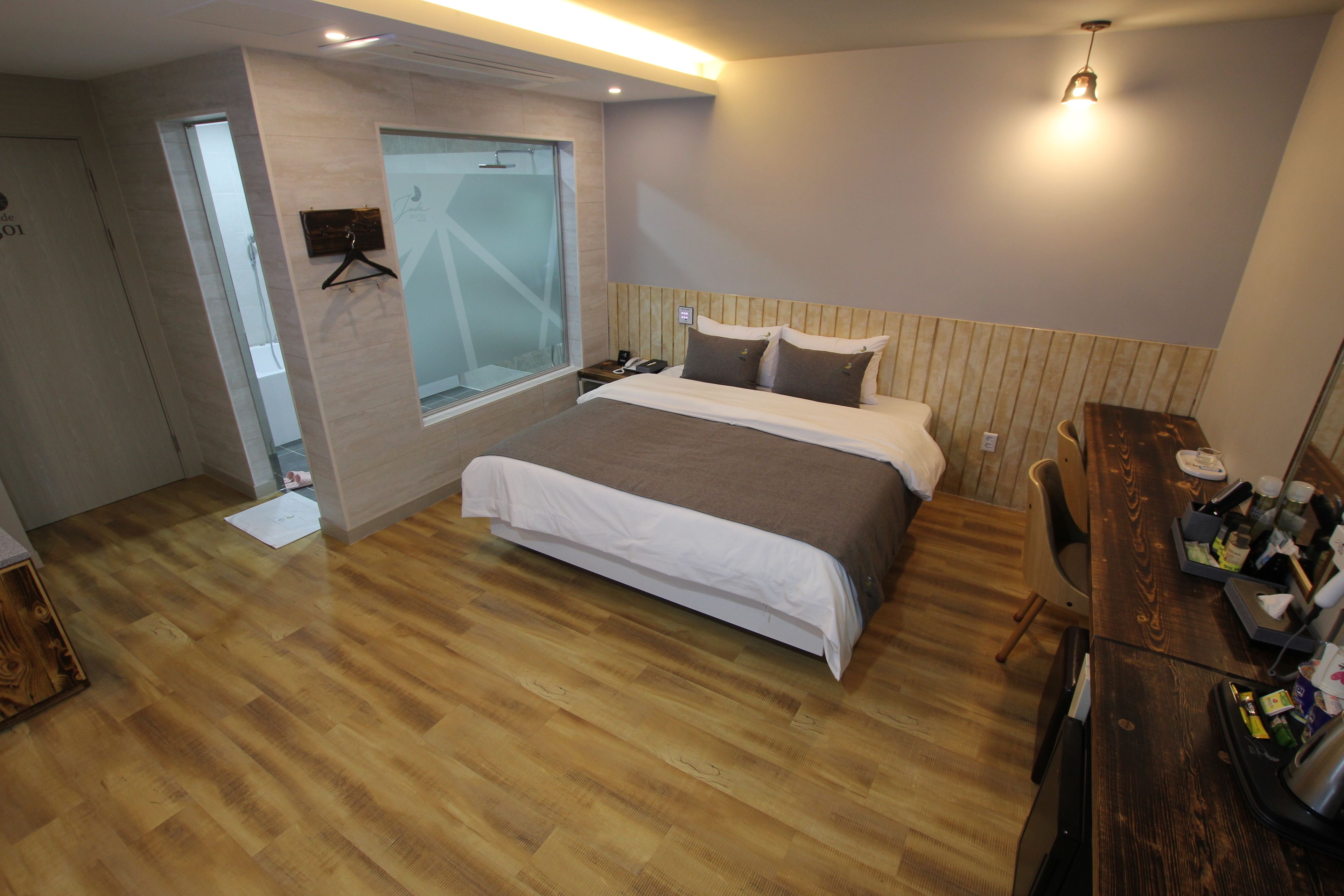 A bedroom with a wooden floor, a bed with a white and brown duvet, a wooden headboard, a bedside table with a lamp, and a window with a view of the outside.