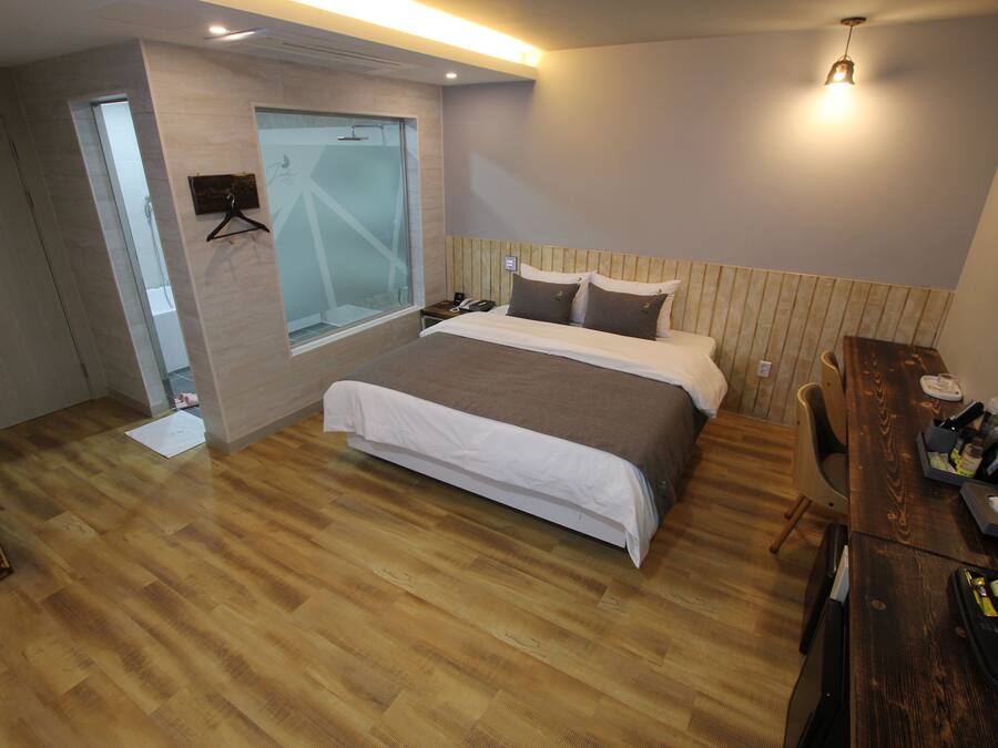 Un dormitorio con piso de madera, una cama con cubrecama blanco y marrón, cabecera de madera, mesita de noche con lámpara y una ventana con vista al exterior.