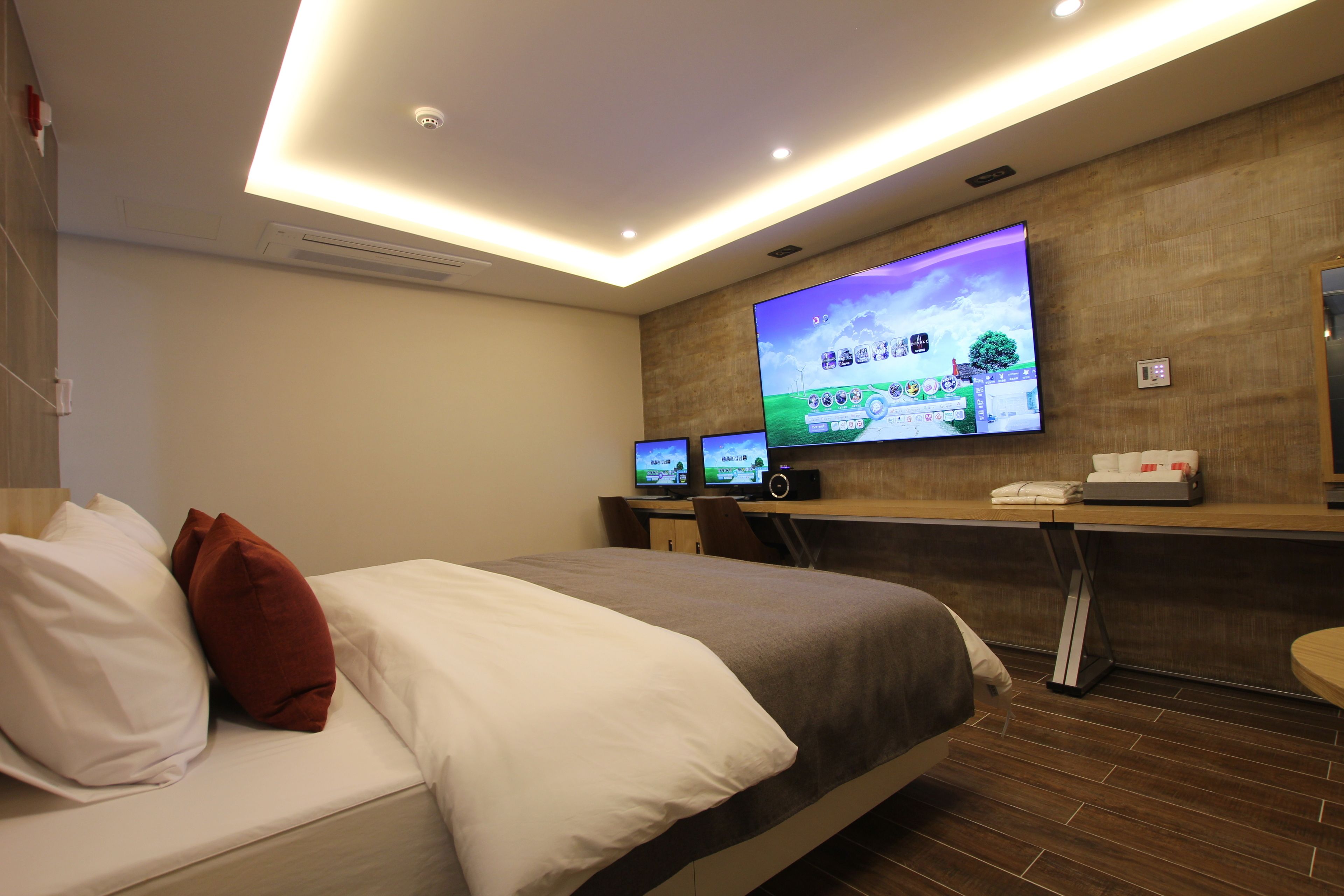 A hotel room with a large bed, a desk with two monitors, and a flat-screen TV mounted on the wall.