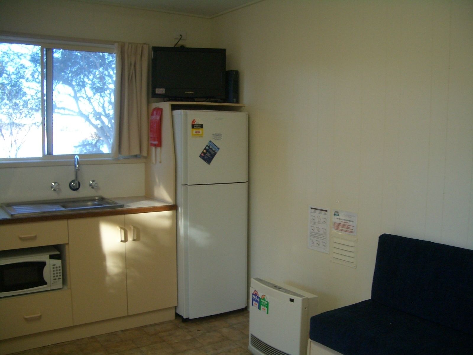 A compact kitchen with a window, a refrigerator, a microwave, a small air conditioner, and a dark-colored couch.