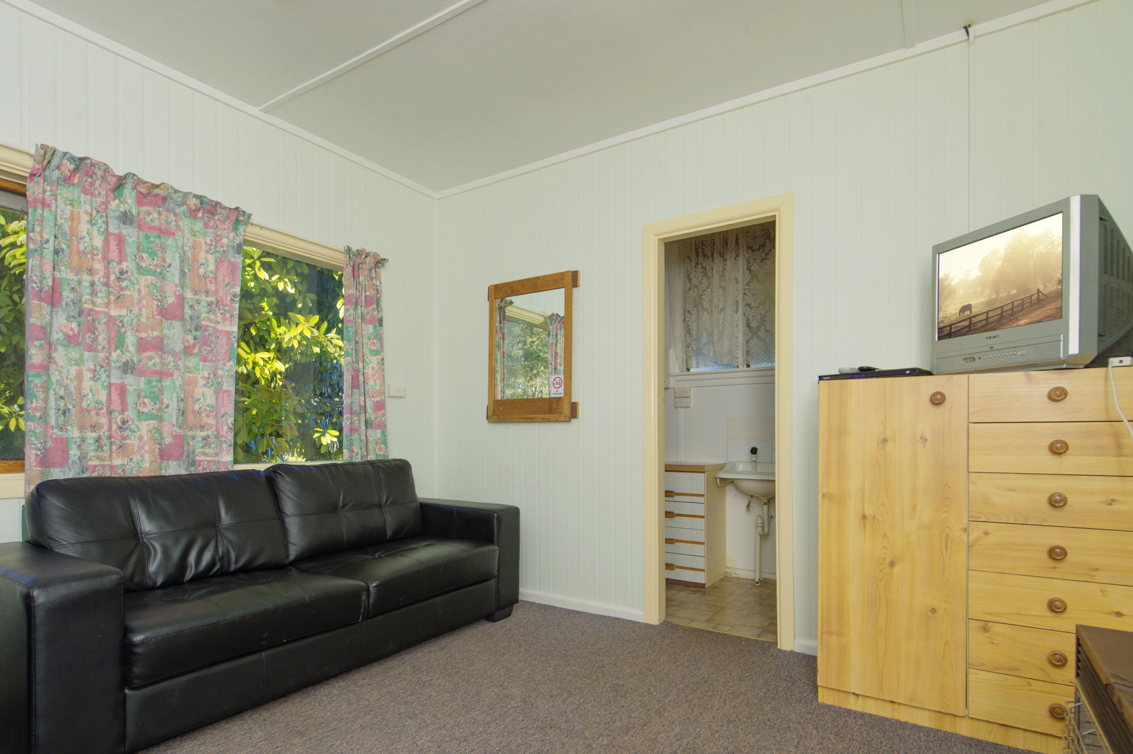 A room with a black leather sofa, a wooden dresser with drawers, a television on top, and a window with floral curtains.