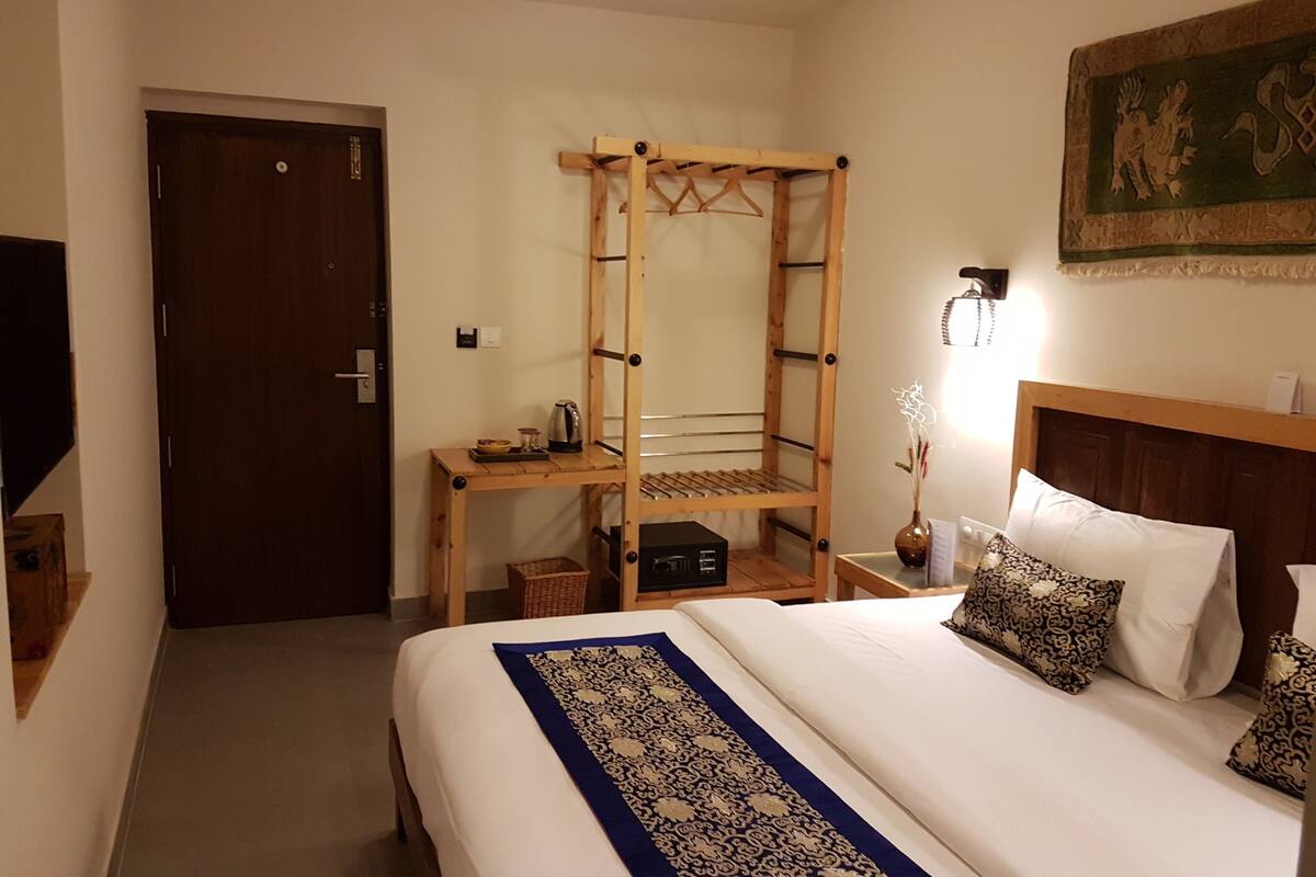A hotel room with a bed, a wooden ladder shelf, a desk with a kettle, and a wall-mounted lamp.